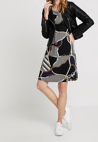 Svart klänning med flerfärgade kedje- och mönsterdetaljer, kombinerad med en svart läderjacka och vita sneakers. Slät tyg.