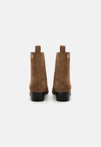 Stivali alla caviglia in suede marrone con una texture liscia, suole in gomma nera e un accento in oro del logo sul tallone. Presentano due linguette per facilitare la calzata.
