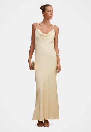 Femeie purtând o rochie tip slip din satin crem cu bretele subțiri, ținând o poșetă mică în mâna dreaptă, și purtând cercei cerc și o brățară tip manșetă.