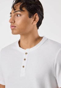 Valkoinen, ribbineulottu lyhythihainen henley-paita, jossa on pyöreä pääntie ja kolme ruskeaa nappikoristetta edessä. Pehmeä rakenne, istuva malli.