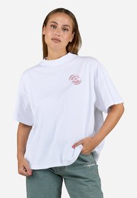 T-shirt blanc surdimensionné avec un col rond et des manches courtes. Présente un petit logo rouge sur la poitrine. Le tissu semble lisse et doux.