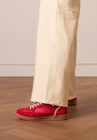 Rote Wildleder- und synthetische Low-Top-Sneaker mit weißen Schnürsenkeln und Gummisohle; getragen mit beigen weit geschnittenen Hosen. Glatte Textur, sportliches Design.