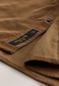 Gros plan d'un tissu en velours côtelé marron avec une étiquette noire portant la mention « Durable Goods Lot 002 » et un détail de bouton-pression.