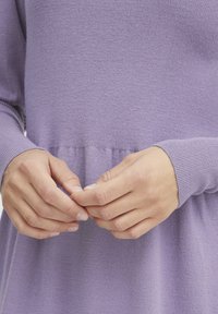 Ljuslila tröja med ribbat mönster och långa ärmar, med en samlad detalj vid midjan, händer som försiktigt håller i tyget.