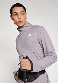 Μακρυμάνικο αθλητικό τοπ σε ανοιχτό μοβ χρώμα με φερμουάρ στον γιακά, με το λογότυπο της The North Face. Το μοντέλο κρατάει μπαστούνια πεζοπορίας και φοράει μαύρο τσαντάκι μέσης.