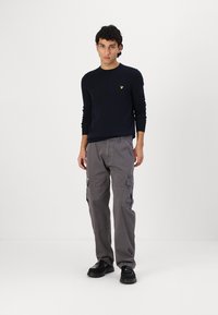 Námornícky pletený sveter s okrúhlym výstrihom, šedé cargo nohavice s bočnými vreckami a čierne topánky, vytvárajúce neformálny outfit.