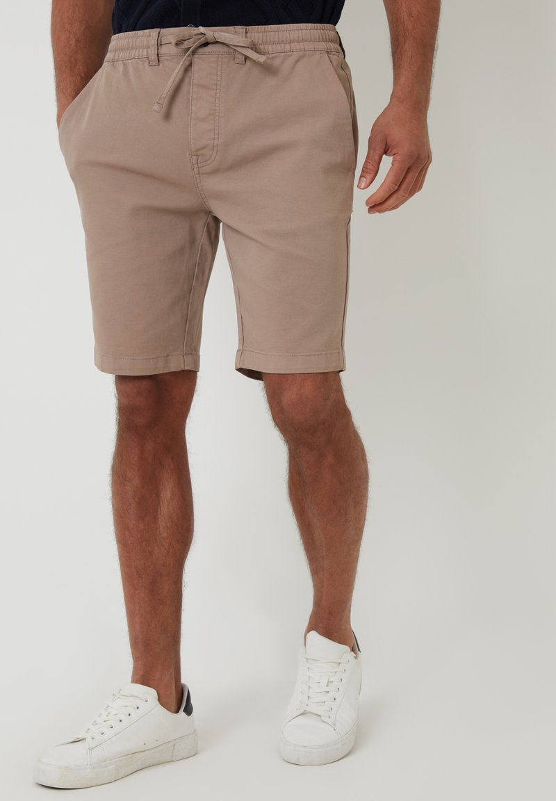 Shorts en coton beige avec une taille élastique et un cordon de serrage. Comprend des poches latérales et un design ajusté, associés à des sneakers blanches.