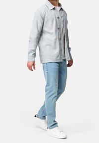 Camicia grigia chiaro con colletto, indossata sopra una t-shirt bianca. Abbinata a jeans blu chiaro e sneaker bianche. Design casual.