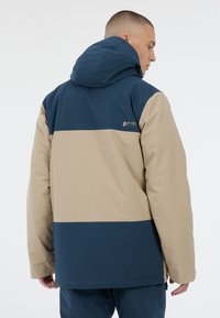 Herren Kapuzenjacke mit einem Farbschnitt-Design in Beige und Marineblau, aus glattem, wasserabweisendem Material gefertigt, mit einem Logo auf der Rückseite.