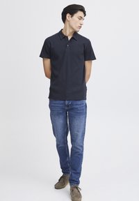 Mørkeblå polo skjorte med korte ermer og ribbet tekstur, med en knappestolpe med to knapper. Brukt sammen med lyseblå denimjeans og brune sko.