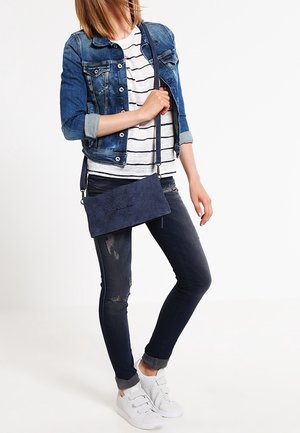 Mørkeblå ruskinds crossbody-taske med rektangulær form, justerbar rem og dekorativ syning; båret med en denimjakke og en stribet skjorte.