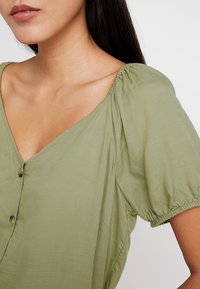 Blusa verde oliva con mangas cortas abullonadas, escote en V y detalle de botones en la parte delantera, hecha de una tela ligera y texturada.