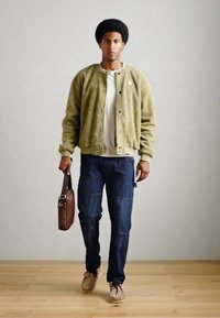 Giacca bomber verde e texture sopra una camicia bianca, abbinata a jeans blu scuri e scarpe marroni. Borsa a tracolla marrone in mano. Sfondo di pavimento in legno.
