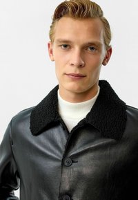 Zwarte leren jas met een gestructureerde zwarte schapenvacht kraag, voorzien van een knoopsluiting en een gladde afwerking. Model draagt er een witte coltrui onder.