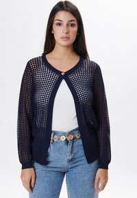 Navyblaue Strickjacke mit offenem Mesh-Design, Rundhalsausschnitt und einem einzelnen goldenen Knopfverschluss. Lange Ärmel mit gerippten Bündchen.