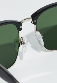 Ochelari de soare cu ramă neagră și lentile de culoare verde închis. Caracteristicile includ accente metalice de tonalitate aurie și pernuțe nazale transparente. Formă pătrată cu detalii texturate.