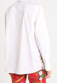 Vit skjorta med knappar av bomull, med en enkel krage och rak passform. Anmärkningsvärda detaljer inkluderar en liten logotyp på baksidan.