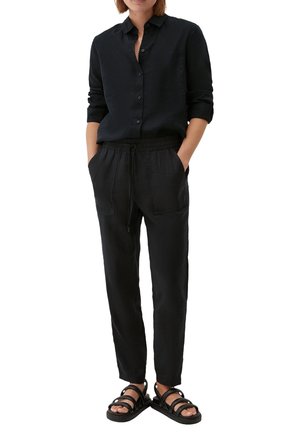 Person iført en sort skjorte med knapper og op-rullede ærmer, sorte bukser med snor og lommer samt sorte sandaler med stropper på hvid baggrund.