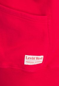 Sudadera roja con una superficie texturizada, que presenta un bolsillo tipo parche y una etiqueta blanca con la inscripción "Levi's Red" en rojo.