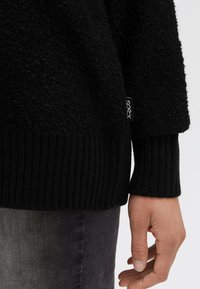 Nahaufnahme einer Hand neben dem Saum eines schwarzen strukturierten Pullis mit Rippbündchen und einem kleinen Stoffetikett.