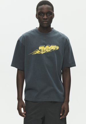 Jeune personne portant un T-shirt ample sombre avec un « W » jaune et un motif de flamme sur la poitrine, debout devant un fond clair uni.