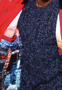 Doudoune rouge avec un motif graphique de paysage urbain et des accents noirs, associée à un pull en tricot texturé bleu marine avec un col rond.