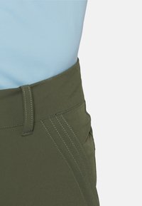 Nahaufnahme der Taille einer olivgrünen Hose mit aufgenähten Nahtdetails, passender Gürtelschlaufe und hellblauem Stoff, der innen eingesteckt ist.