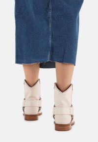 Botines de color beige claro con un cuello en forma de V, correas laterales con herrajes metálicos y un tacón de madera, combinados con una falda de mezclilla azul.