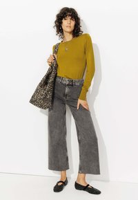 Haut à manches longues vert olive, jean large gris taille haute, ballerines noires avec des clous, et un sac fourre-tout à motifs léopard. Fond blanc.