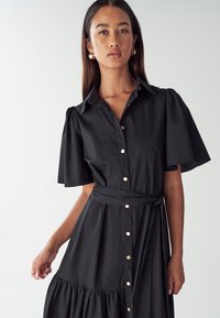 Čierne košeľové šaty s krátkymi nadýchanými rukávmi, viazaným pásom a tierovými volánmi na leme; obsahujú zlaté gombíky a hladký povrch.