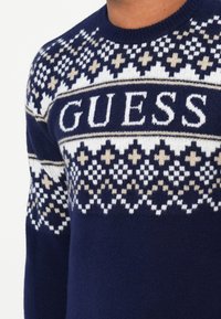 Maglione blu navy con un motivo a maglia texturizzata. Accenti geometrici bianchi e beige con un prominente logo "GUESS" sul petto.