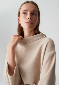 Giovane donna con capelli castano chiaro, che indossa un maglione beige a coste, che tiene delicatamente il colletto del maglione, guardando verso l'alto su uno sfondo neutro.