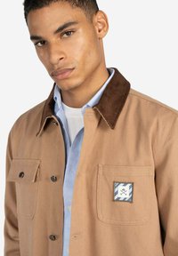 Chaqueta de lona color arena con un cuello de pana marrón, dos bolsillos en el pecho y un detalle de parche azul y blanco. El modelo lleva puesta una camisa azul clara debajo.