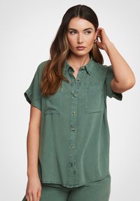 Camicia verde a maniche corte con bottoni in tessuto morbido, dotata di colletto, due tasche sul petto e una vestibilità morbida. Cuciture a vista in evidenza.