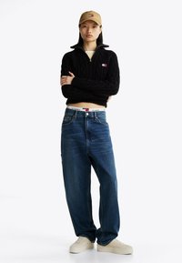 Maglione nero a maglia grossa con zip, jeans blu a gamba larga, cappello beige e scarpe da ginnastica bianche. Dettagli notevoli includono accenti del logo sul maglione e sulla fascia della vita.