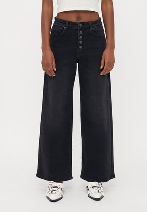 Femme portant un jean noir taille haute à jambes larges avec braguette à boutons et chaussures blanches à brides, debout devant un fond clair uni.