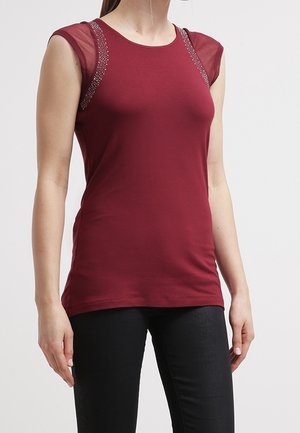 Frau trägt ein ärmelloses, dunkelrotes Oberteil mit transparenten Mesh-Akzenten und Verzierungen auf den Schultern, kombiniert mit einer schwarzen Hose.