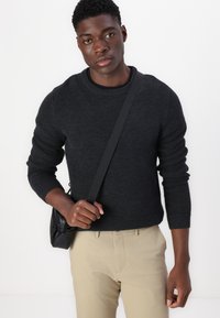 Pull en maille noire à col rond et poignets côtelés, associé à un pantalon beige. Le mannequin porte un petit sac noir en bandoulière.