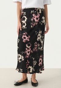 Jupe noire à motif floral avec de grandes fleurs roses et crème, au design fluide, en tissu léger et avec un ourlet évasé, portée avec des chaussures plates noires.