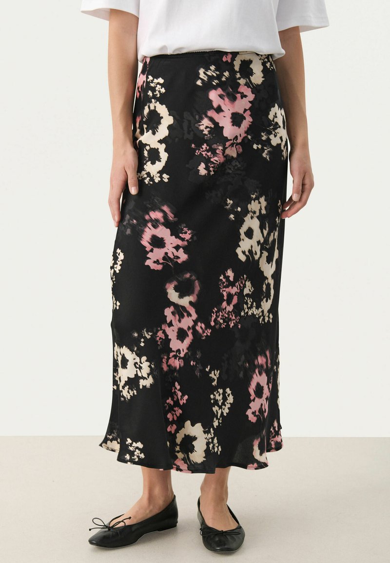 Jupe noire à motif floral avec de grandes fleurs roses et crème, au design fluide, en tissu léger et avec un ourlet évasé, portée avec des chaussures plates noires.