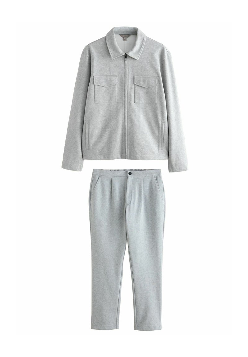 Giacca grigia con zip, due tasche frontali e colletto, abbinata a pantaloni grigi coordinati con chiusura a bottone e pieghe frontali.