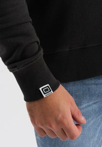 Felpa nera con polsini a costine, che presenta un'etichetta bianca con la scritta "ombre". Primo piano del polso e della mano contro un tessuto di denim.