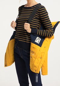 Pull rayé bleu marine et jaune à col rond, assorti d'une veste matelassée jaune et bleu marine, avec un écusson et des accessoires contrastants.