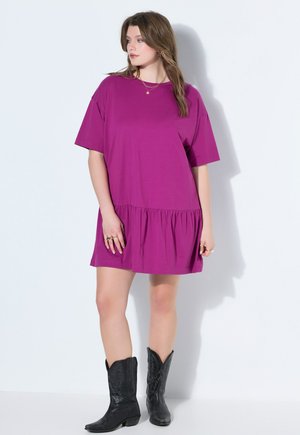 Frau trägt ein lockeres lila kurzärmeliges Kleid und schwarze Cowboy-Stiefel, steht vor einer weißen Wand und schaut zur Seite.