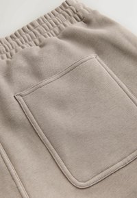 Tissu beige clair avec poche arrière rectangulaire cousue et ceinture élastique, montrant la texture et les détails des coutures.