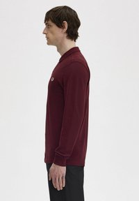 Polo a maniche lunghe bordeaux con colletto classico, logo ricamato sul petto e polsini a costine, abbinato a pantaloni neri.