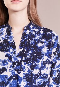Blusa blu e bianca con motivo a fantasia, caratterizzata da un collo a V, tessuto leggero e texture morbida, con un vivace design astratto.