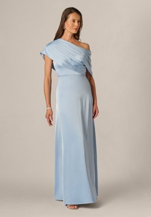 Femme portant une robe bleu clair, asymétrique, longue jusqu'au sol avec des détails plissés, debout devant un fond neutre uni.