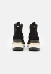 Botas de tobillo de ante negro con un cuello de punto acanalado, que presentan un acento dorado en el tacón y una suela blanca y negra. Textura suave.