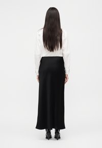 Μακριά μαύρη φούστα από λεία υφή, σε συνδυασμό με λευκό πουκάμισο με λεπτομερείς μανσέτες. Πλάγια όψη από πίσω, φορεμένα με ψηλά μπότες με τακούνι.